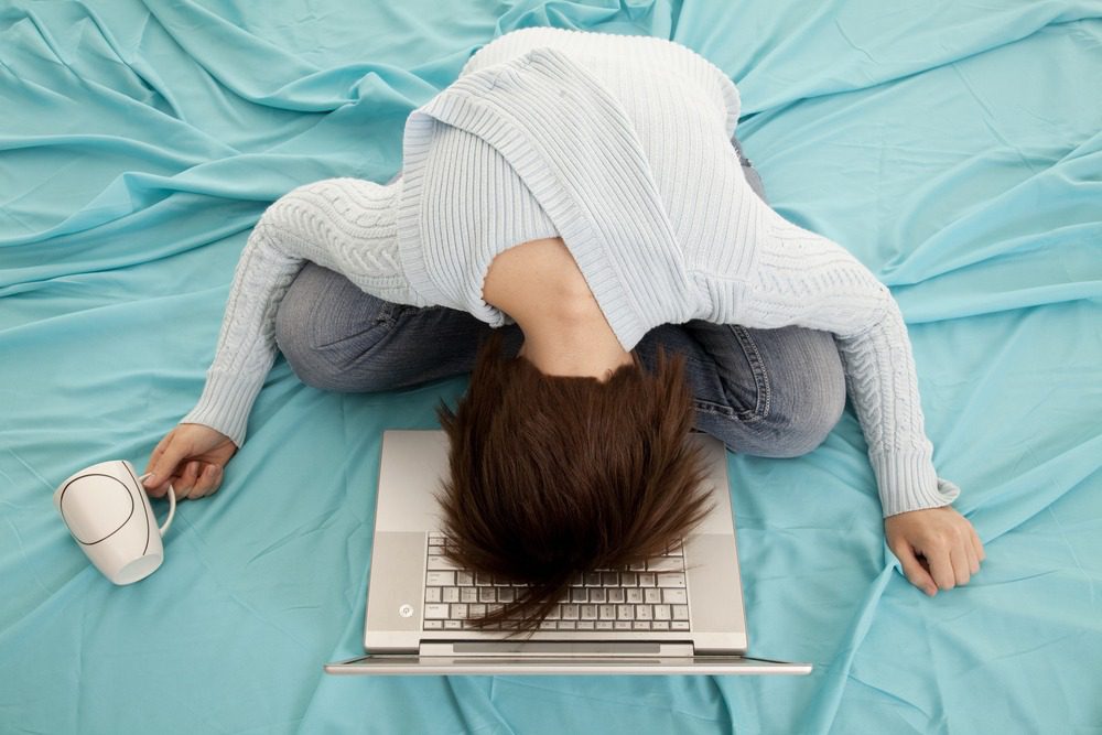 a woman in bed with her head laying on the computer holding her coffee mug.