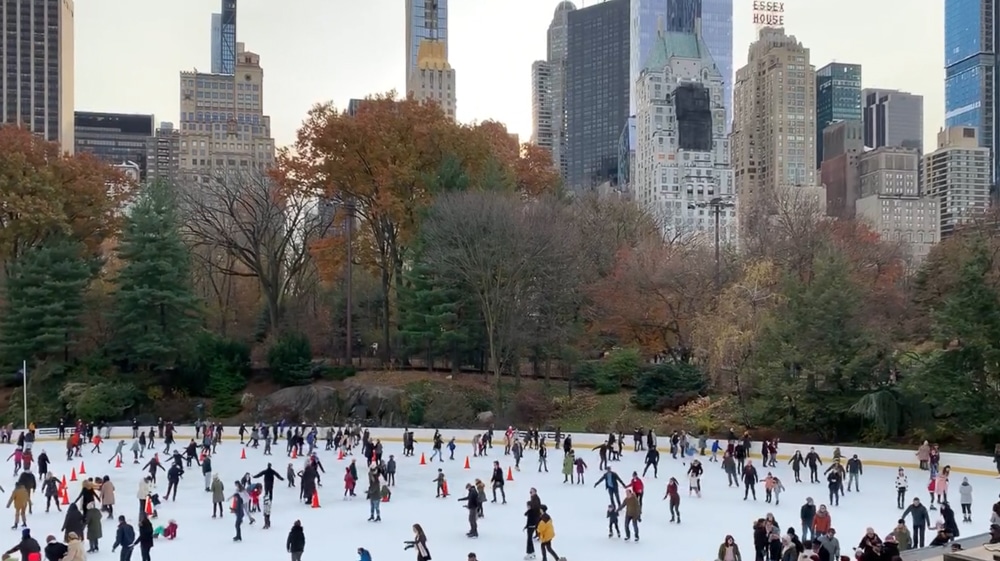 wollman ice skating rink in NYC's central park with trees and skyscrapers in the distance and lots of skaters on the ice