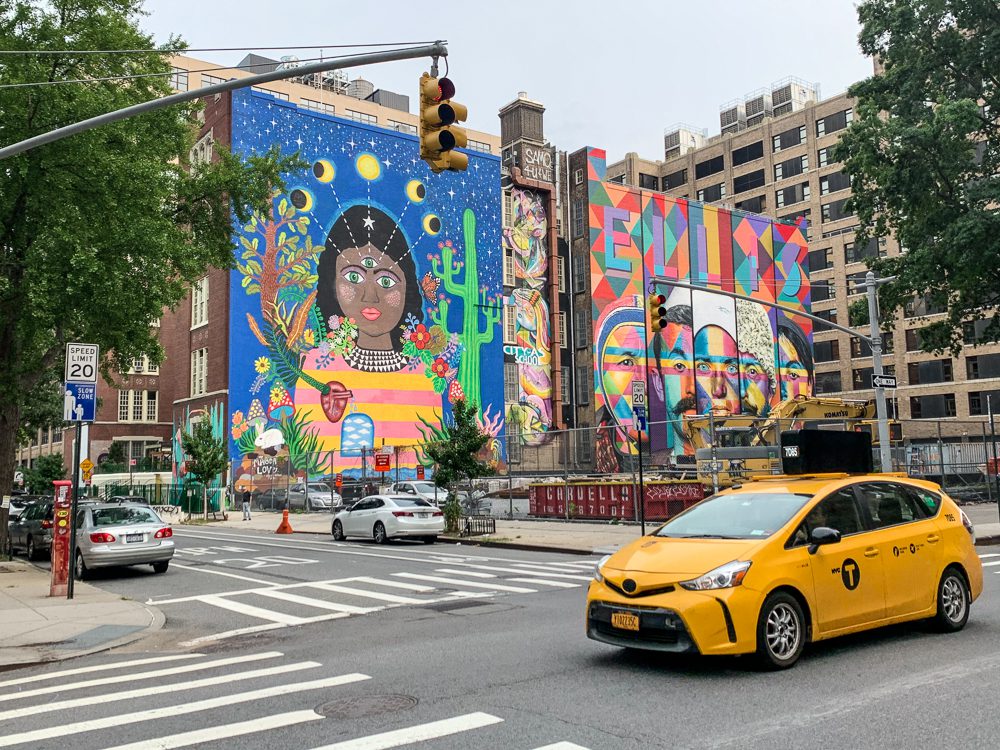 A taxi driving through a NYC intersection with street art murals on the walls of the buildings in the background
