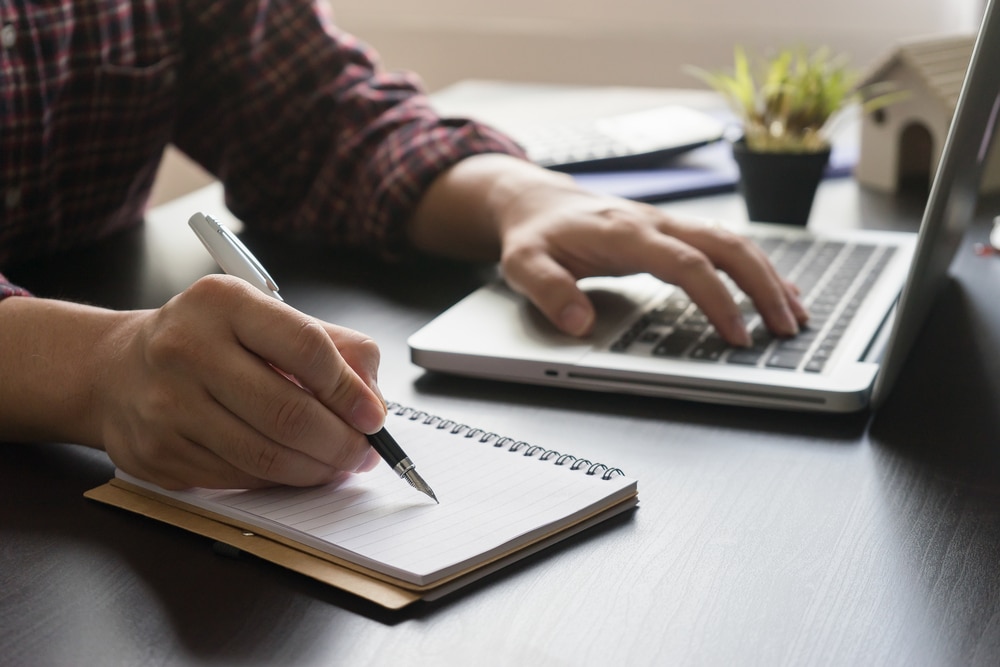Male putting his ideas into writing in a notebook with a laptop nearby